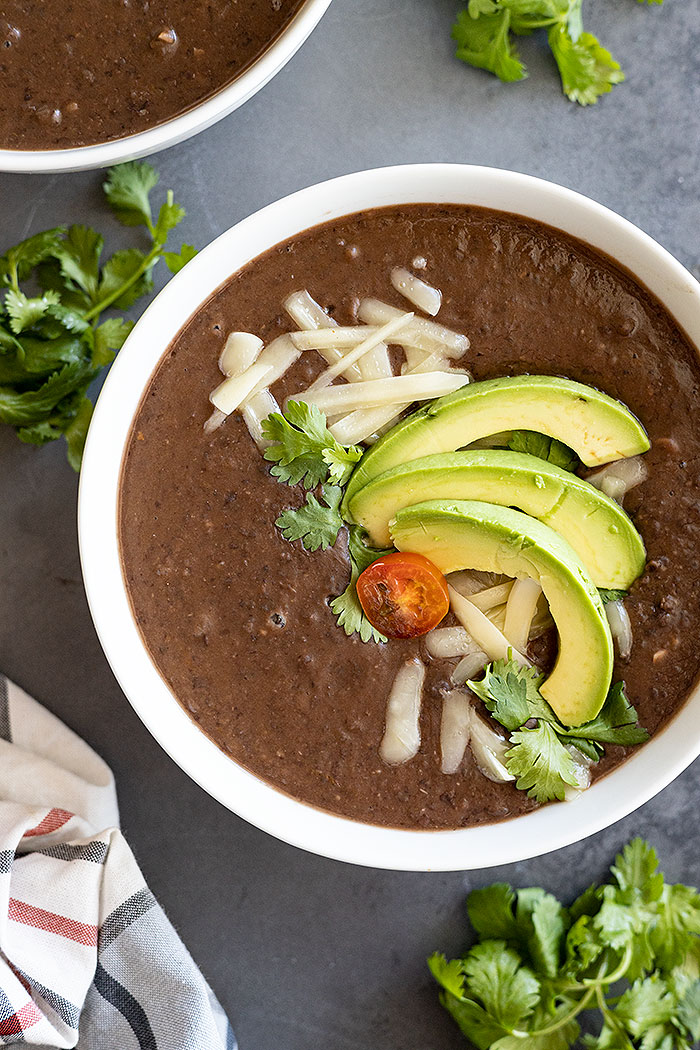 Overhead view of black bean soup in a white bowl garnished with avocado, cheese, cilantro, and tomato. 