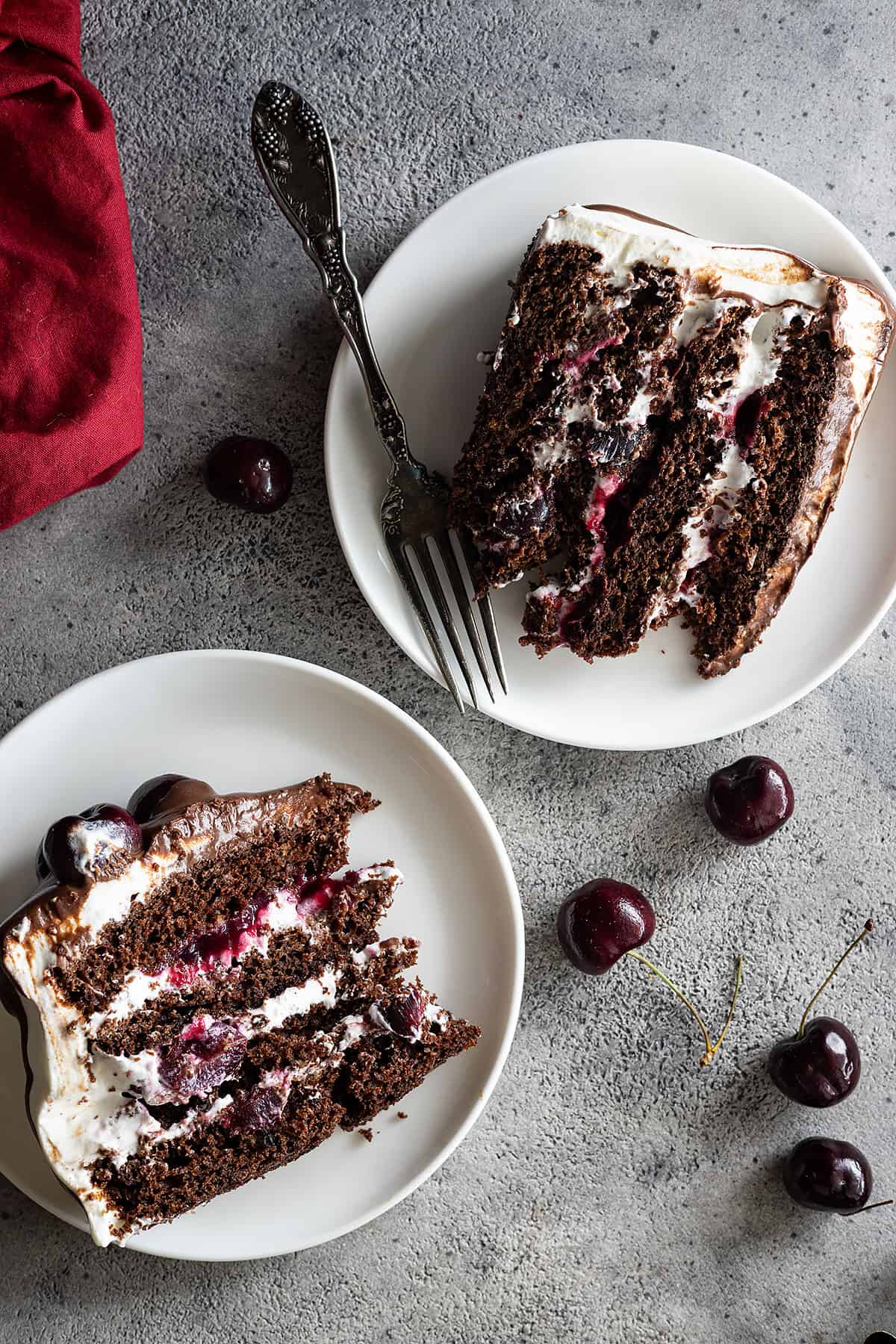 Overhead view of 2 slices of cake on white plates with red napkin off to the side. 