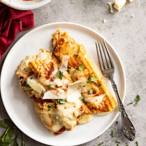 Overhead view of manicotti on a white plate. Garnished with fresh parsley and parmesan cheese. Fork and red napkin off to the side.