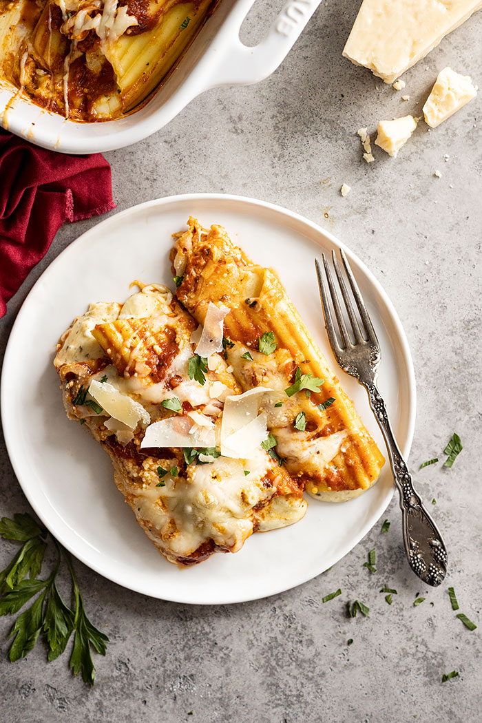 Overhead view of manicotti on a white plate. Garnished with fresh parsley and parmesan cheese. Fork and red napkin off to the side. 
