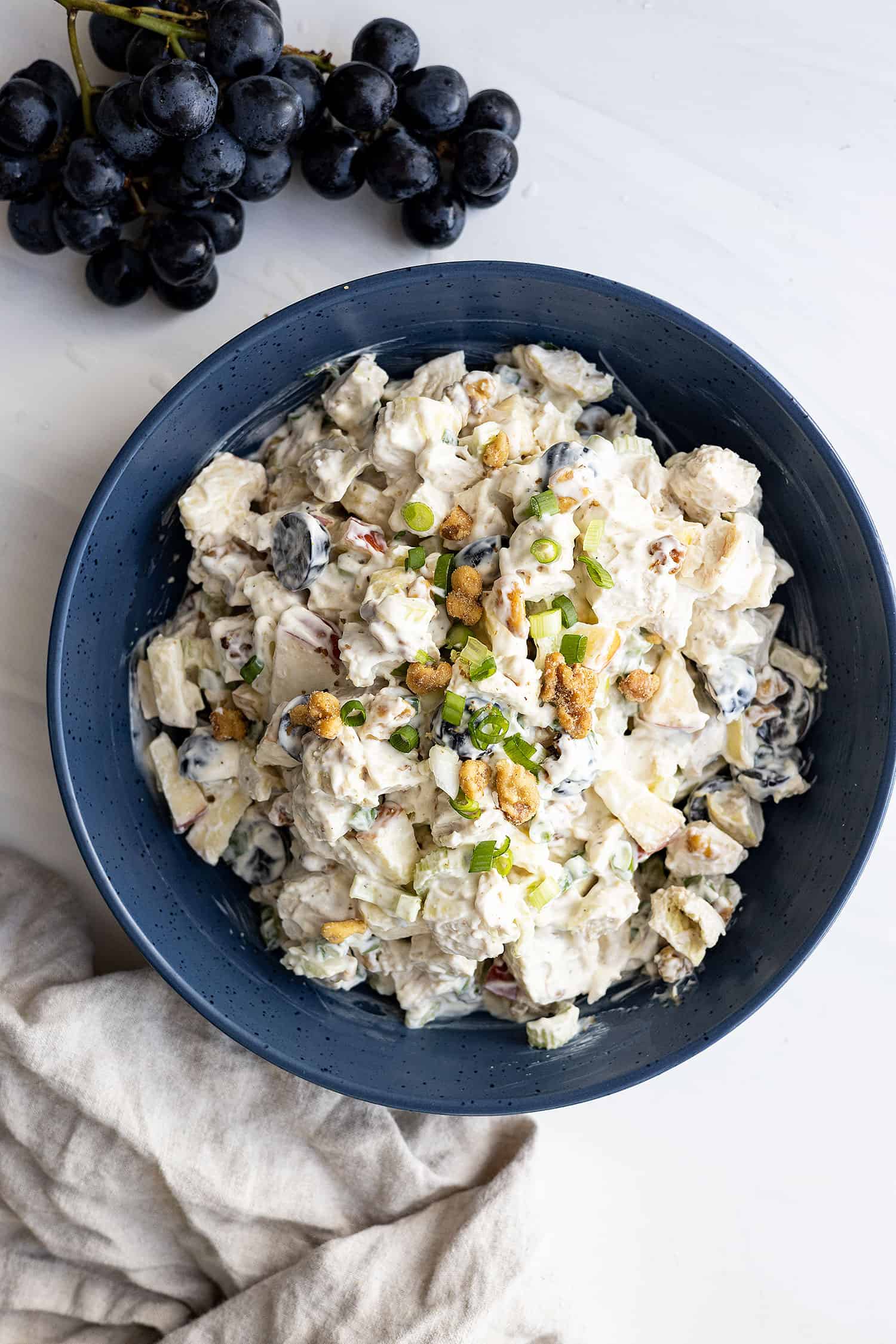 Overhead view of chicken waldorf salad in a large dark blue bowl garnished with chopped walnuts and green onion. Purple grapes in the corner and a light colored napkin beside the bowl. 
