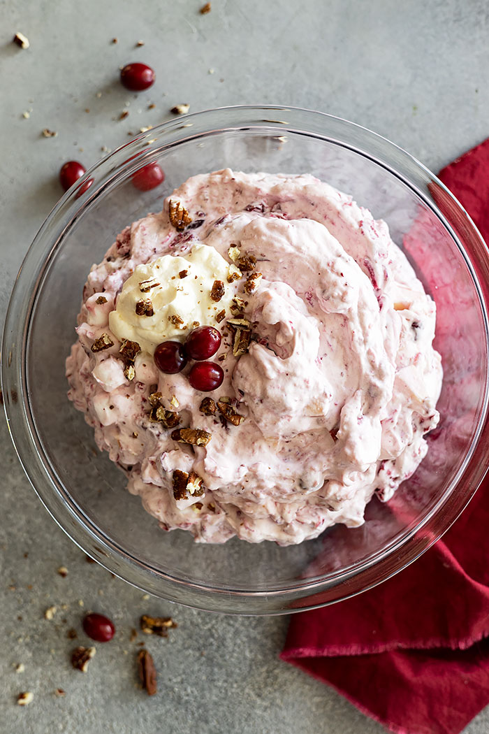 Overhead view of cranberry fluff salad in a bowl garnished with fresh cranberries and pecans. Red napkin off to the side. 
