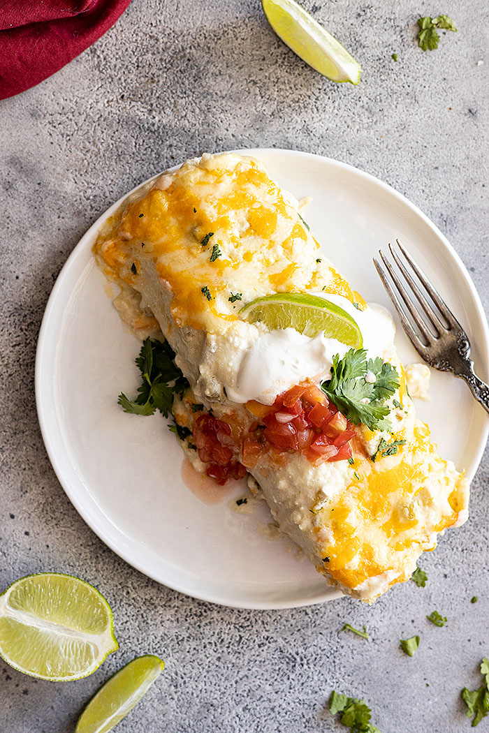 Overhead view of enchiladas served on a white plate garnished with sour cream, cilantro, and lime wedges. 