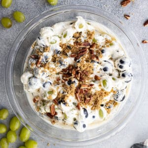 Overhead view of creamy grape salad in a bowl garnished with chopped nuts and brown sugar. Grapes and nuts sprinkled around the bowl.