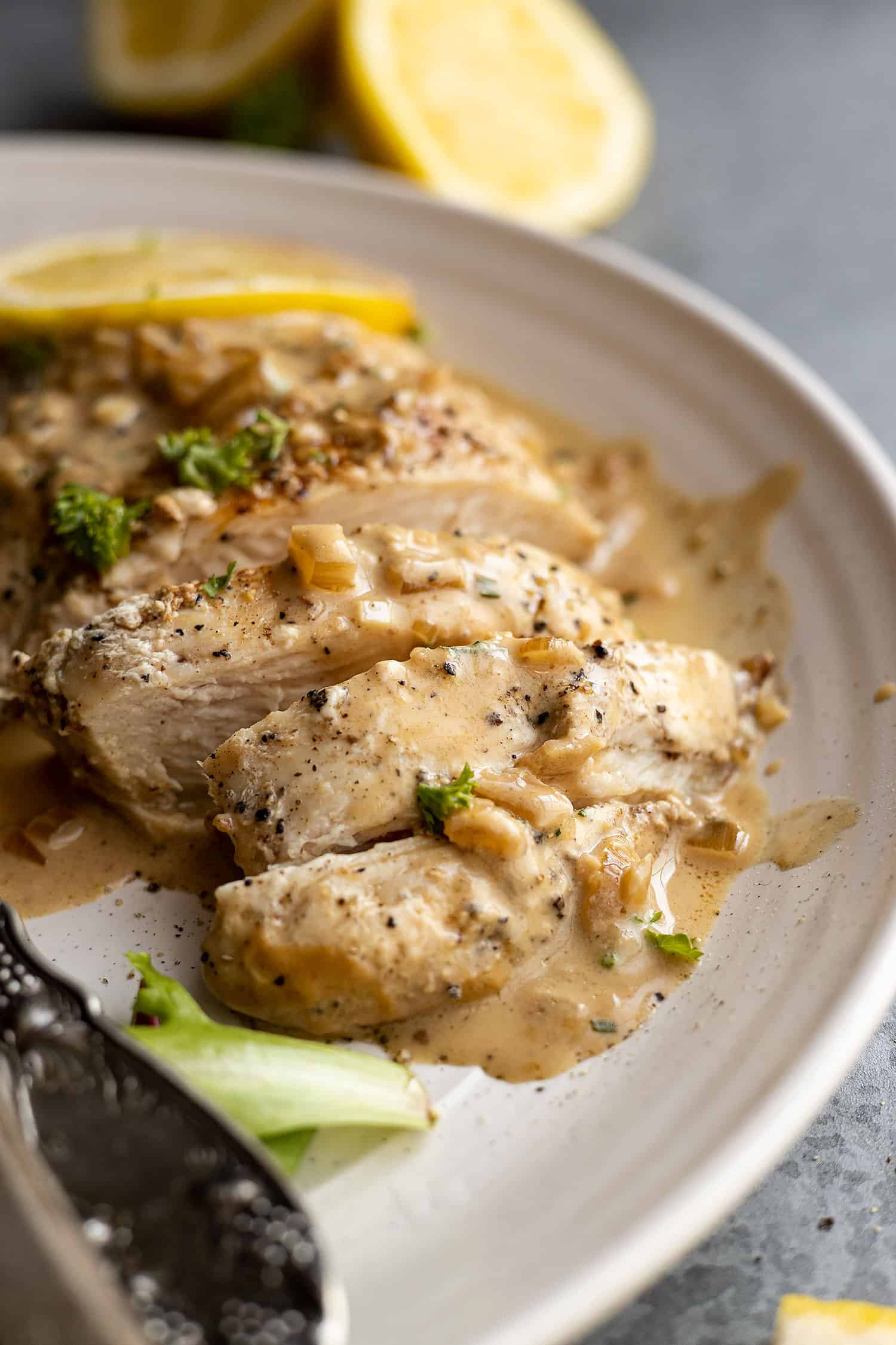 Close up of creamy lemon chicken sliced on a plate with a fresh cut lemon in the background. 