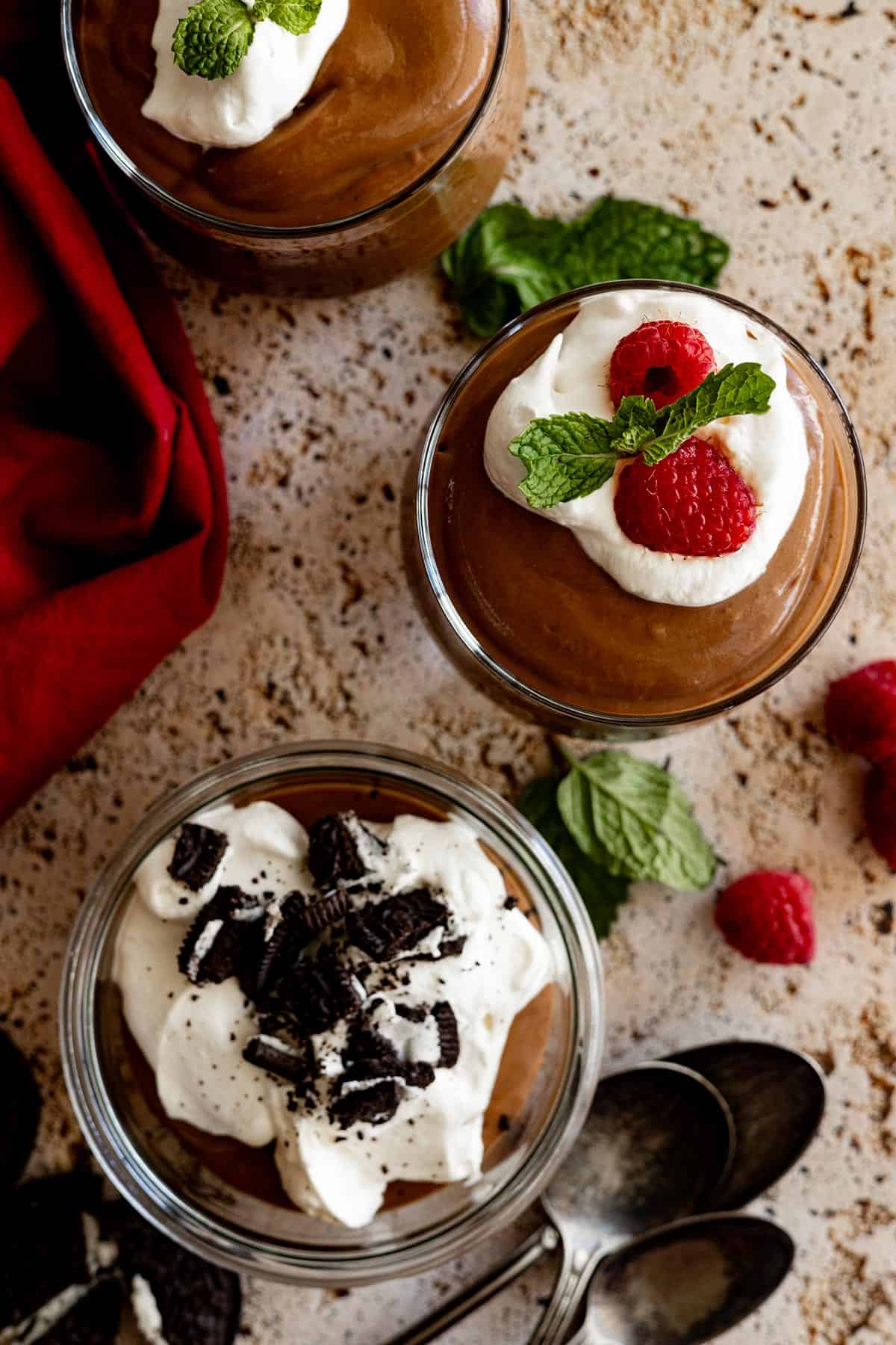 Overhead view of dark chocolate pudding in three cups topped with different toppings. Spoons off to the side. 