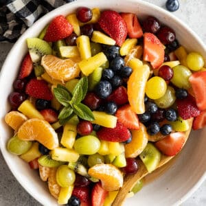 Overhead view of easy fruit salad in a cream bowl with a spoon in the salad. Garnished with mint.