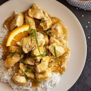 Overhead view of easy orange chicken on a bed of rice and garnished with green onions, sesame seeds, and orange slices.