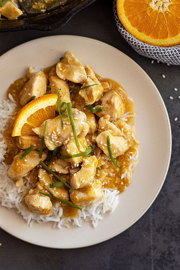 Overhead view of easy orange chicken on a bed of rice and garnished with green onions, sesame seeds, and orange slices.
