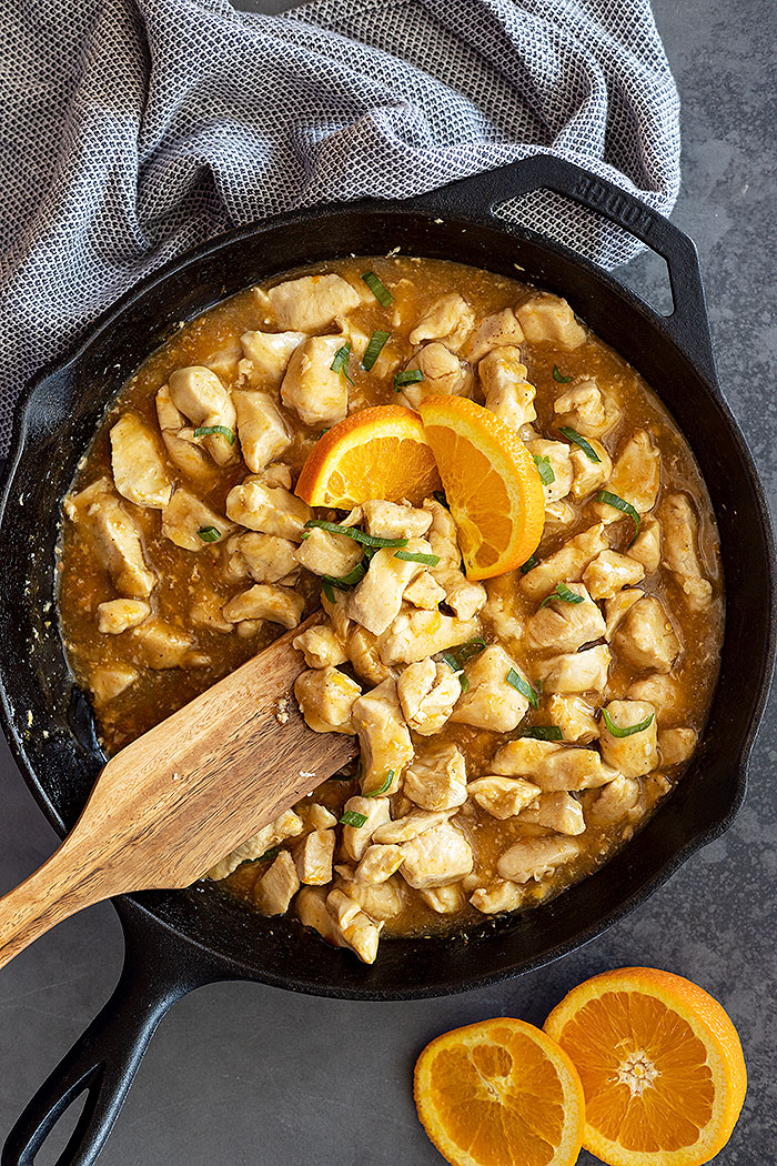 Overhead view of easy orange chicken in a cast iron skillet garnished with green onion and fresh orange wedges.