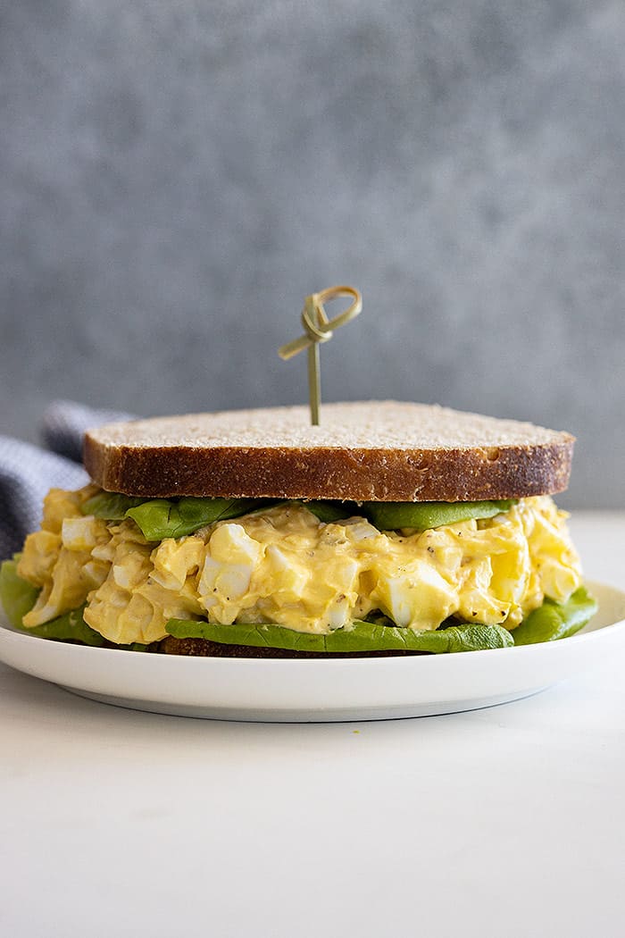 An egg salad sandwich on a plate. Sandwich also has a large lettuce leaf and a decorative toothpick in the center. 
