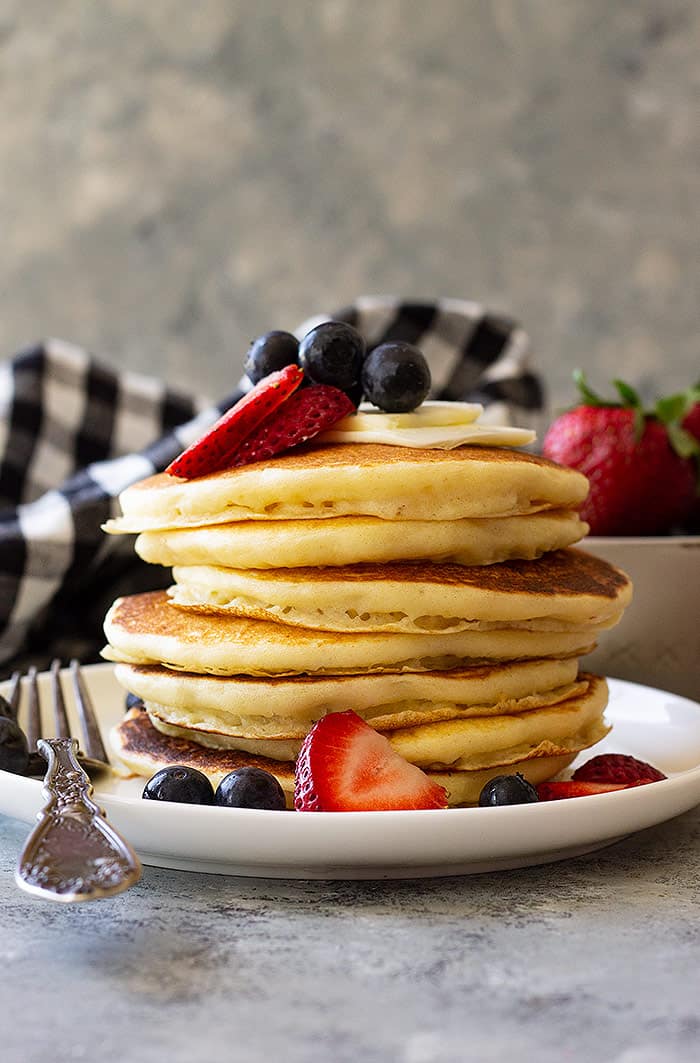 Tall stack of pancakes topped with berries and butter waiting for a splash of syrup.