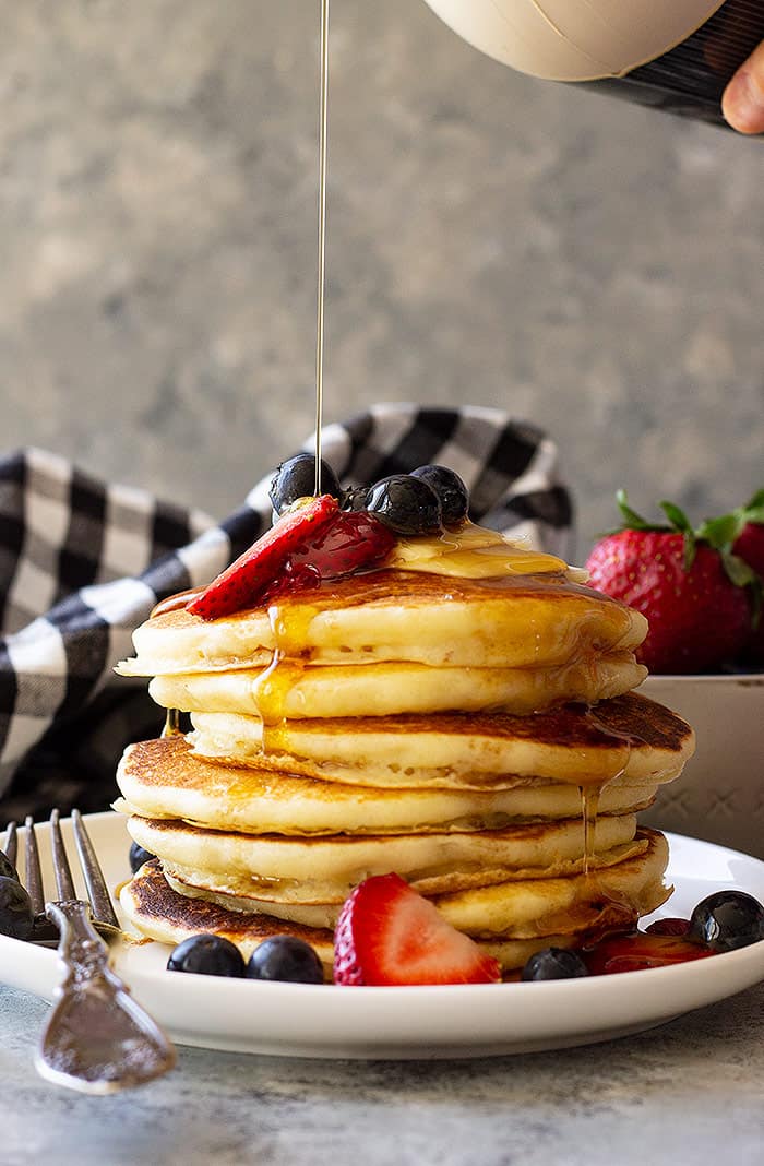 Tall stack of Fluffy Buttermilk Pancakes topped with fresh berries and being drizzled with syrup. 