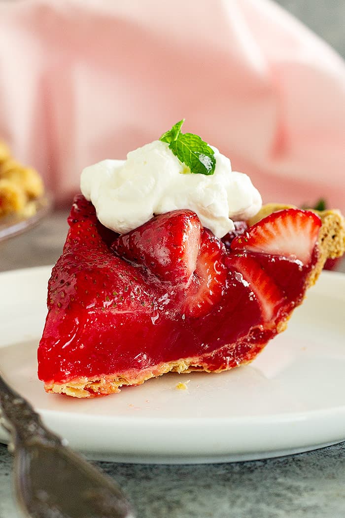 A piece of strawberry pie on a white plate topped with whipped cream and mint.