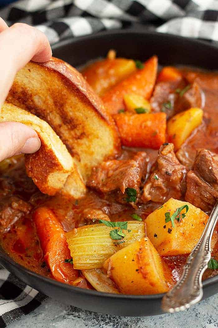 closeup: a hand dunking a piece of bread into a bowl of hearty beef stew with veggies and beef showing