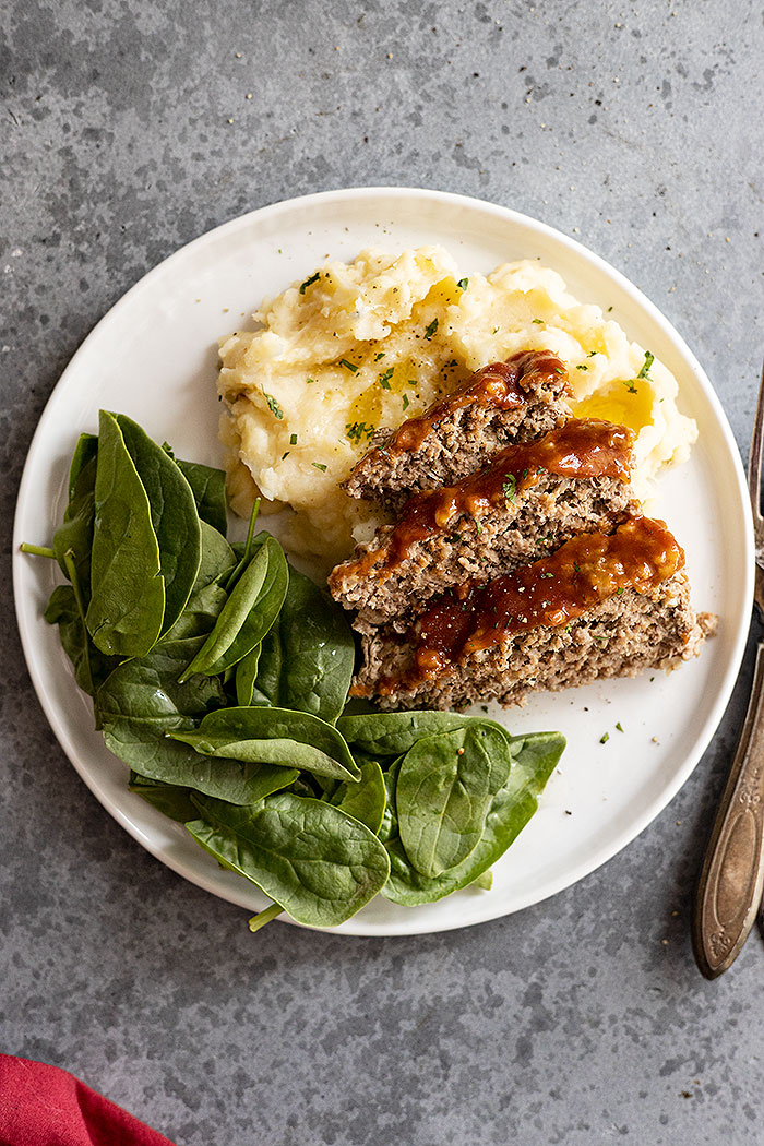 Overhead view of meatloaf and mashed potatoes on a white plate with a fork and red napkin off to the side.