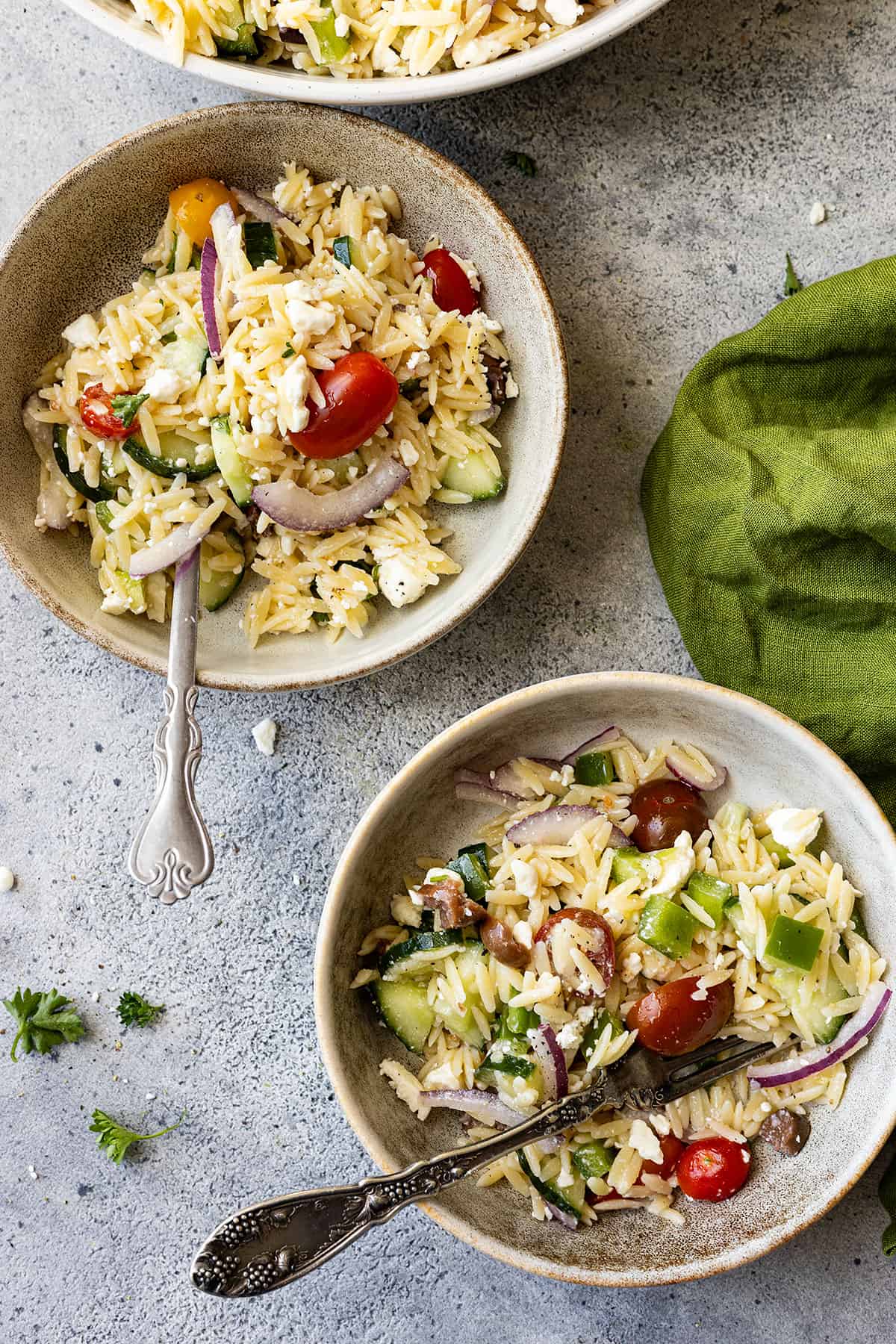 Two bowls with pasta salad and forks in each ready to eat. Green napkin and bits of parsley off to the sides. 