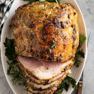 Overhead shot of ham on a white plate garnished with herbs. Part of the ham is sliced.