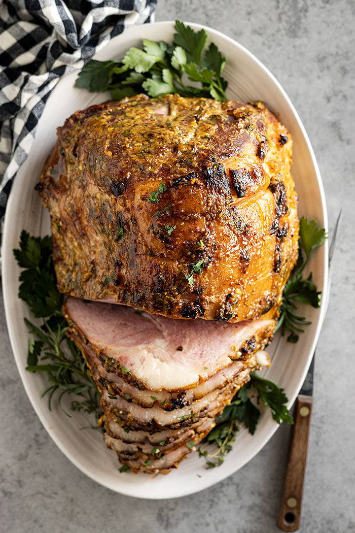 Overhead shot of ham on a white plate garnished with herbs. Part of the ham is sliced. 
