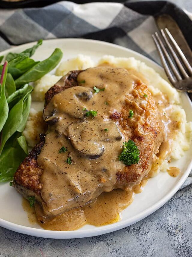 A pork chop on a plate with mash potatoes and topped with a creamy mushroom gravy.