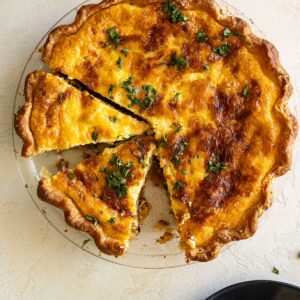 Overhead view of quiche in pie plate with slices cut and one removed and on a plate in the corner.