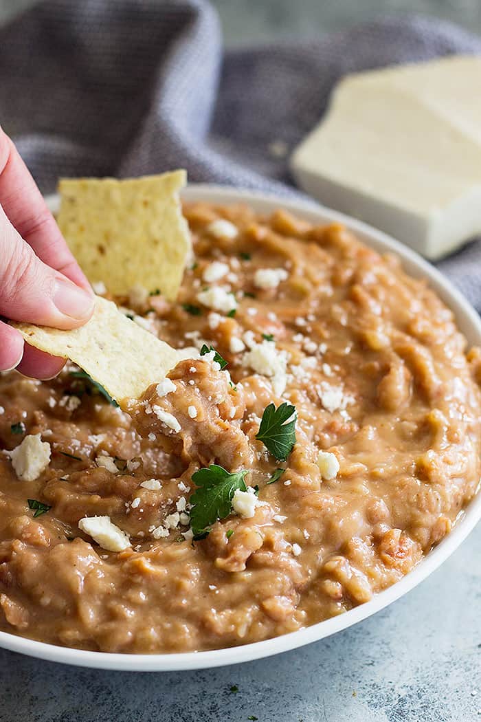 Big bowl of refried beans sprinkled with cheese. A chip is dipping into the beans. 