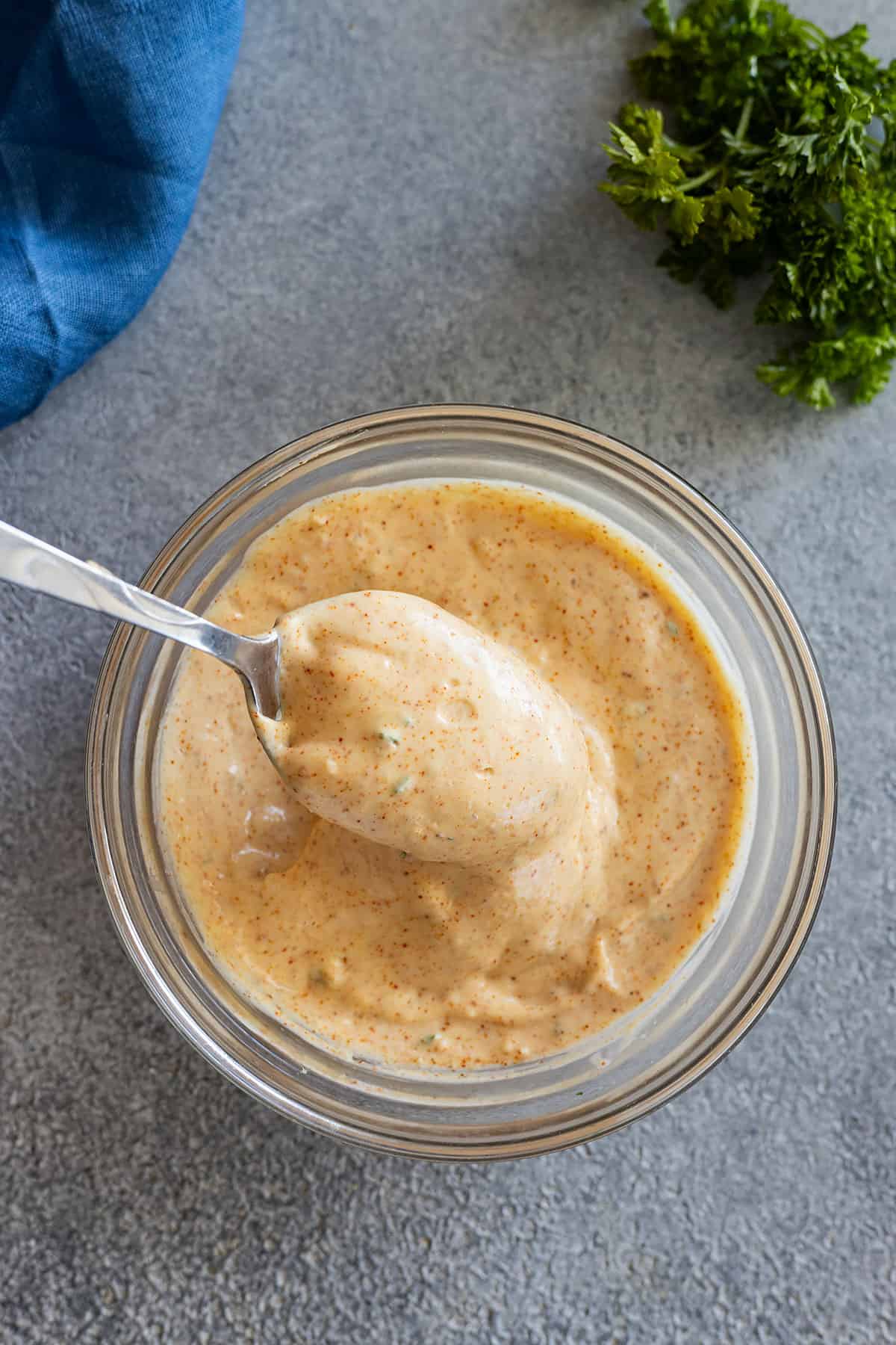 Overhead view of a bowl of remoulade sauce with a spoon dipping into it. Parsley and a blue napkin in the corners.