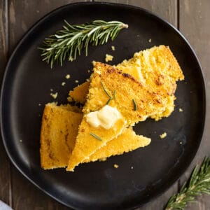 Overhead view of slices stacked on a black plate with melting butter on the top. Rosemary to the side.