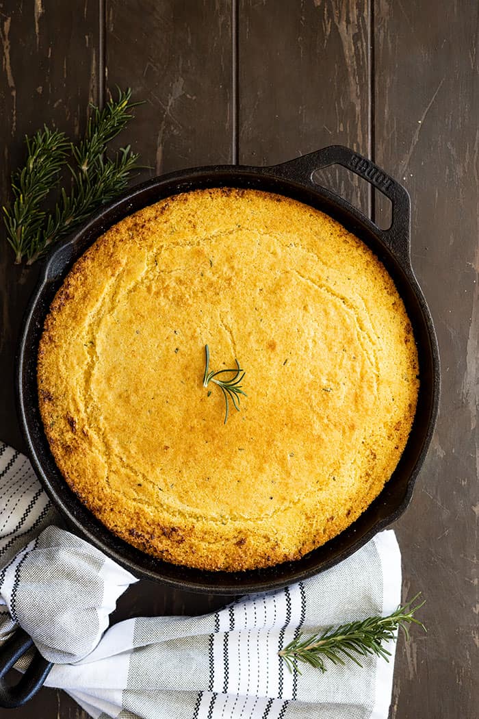 Overhead view of rosemary cornbread fresh from the oven in a cast iron skillet. Rosemary and a towel to the side of the picture.