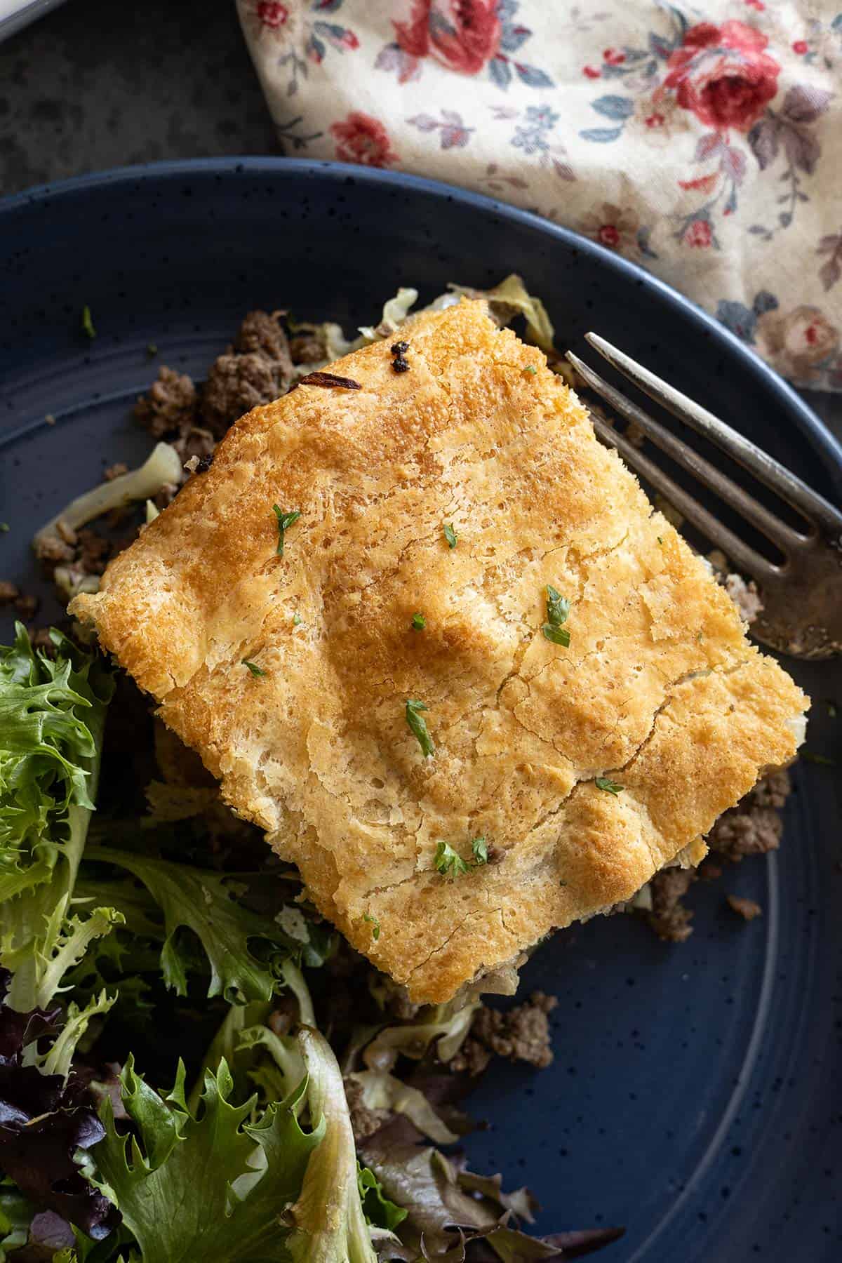 Overhead view of a slice of runza casserole on a dark blue plate with a salad off to the side. Floral napkin in the corner. 