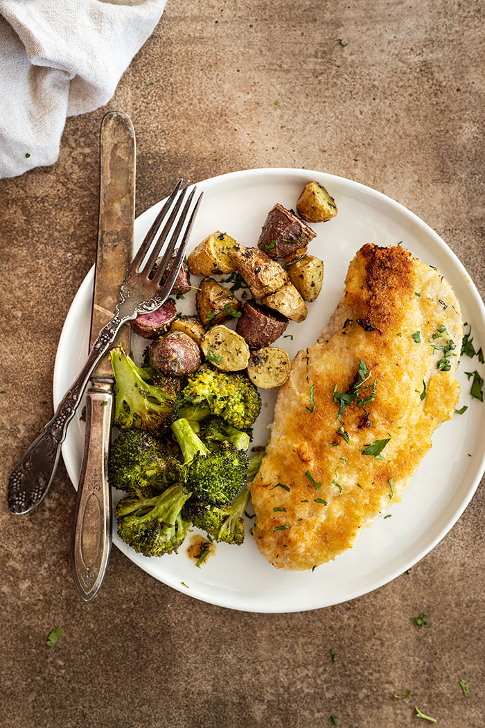Overhead view of a piece of chicken on a white plate with potatoes and broccoli. Fork and knife off to the side. 