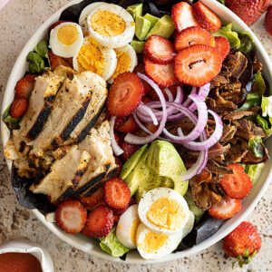 Overhead view of a large bowl of strawberry cobb salad with dressing off to the side.