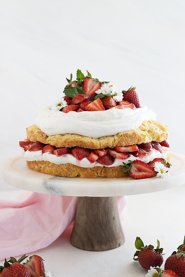 Straight on shot of strawberry shortcake cake on a cake stand and garnished with mint and strawberries cut in half.