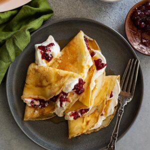 Overhead view of Swedish pancakes on a dark plate. They have whipped cream and lingonberries inside. Fork and green napkin to the side.