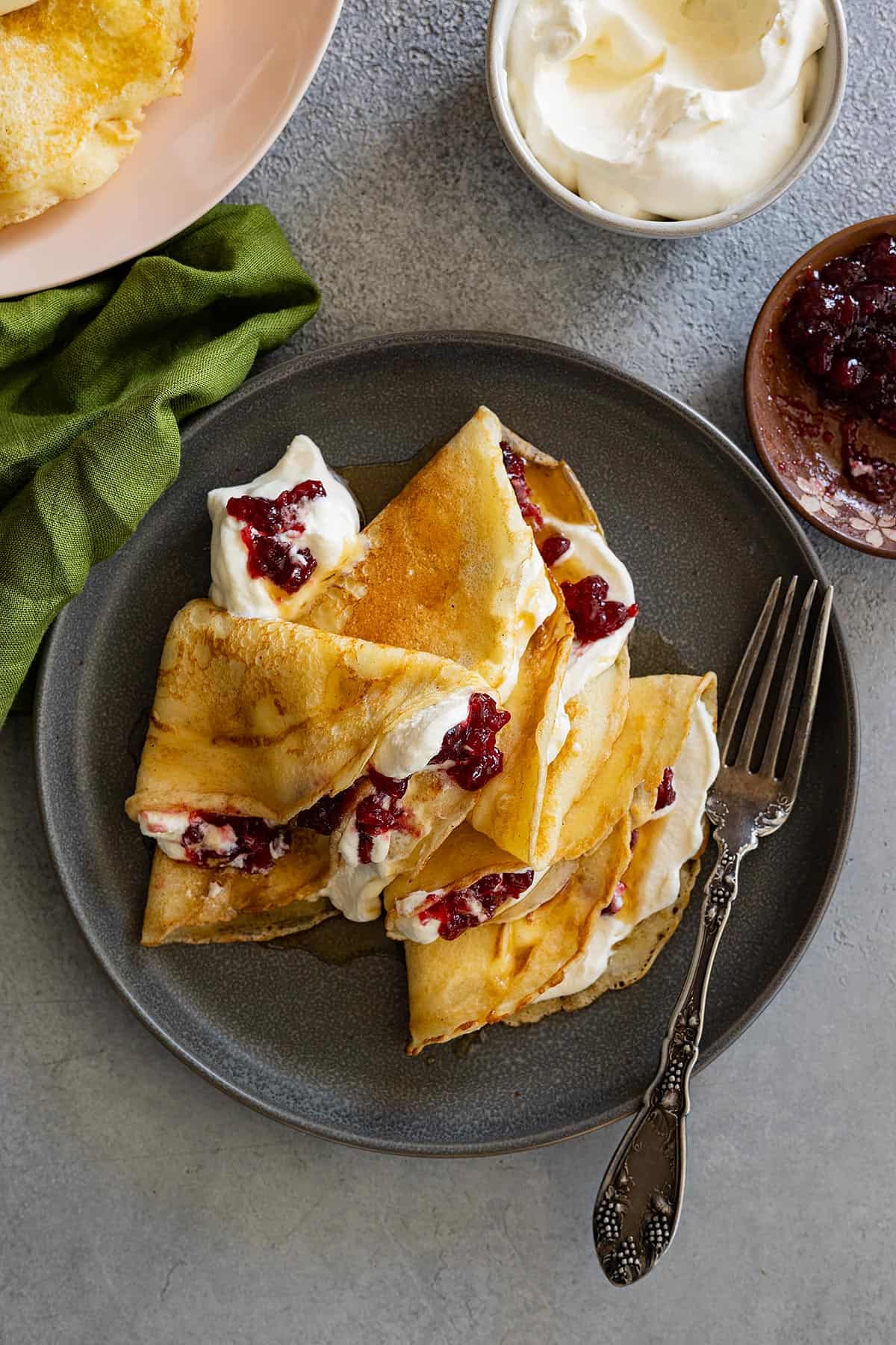 Overhead view of Swedish pancakes on a dark plate. They have whipped cream and lingonberries inside. Fork and green napkin to the side. 