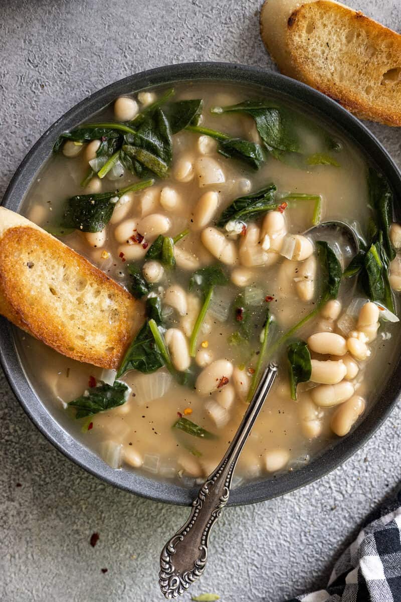 Overhead view of a bowl of tuscan bean soup in a dark bowl. Crusty bread off to the side and a spoon in the soup.