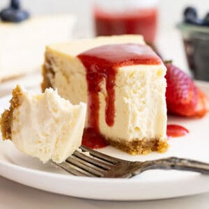 Straight on view of vanilla cheesecake on a white plate, strawberry sauce over the top, and a bite taken out showing how creamy and smooth it is.